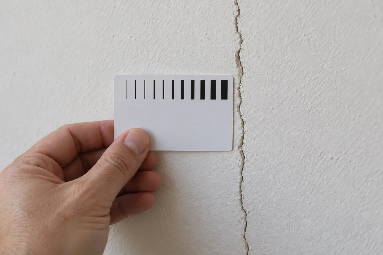 A hand holding a crack-width gauge card against a crack in a painted wall